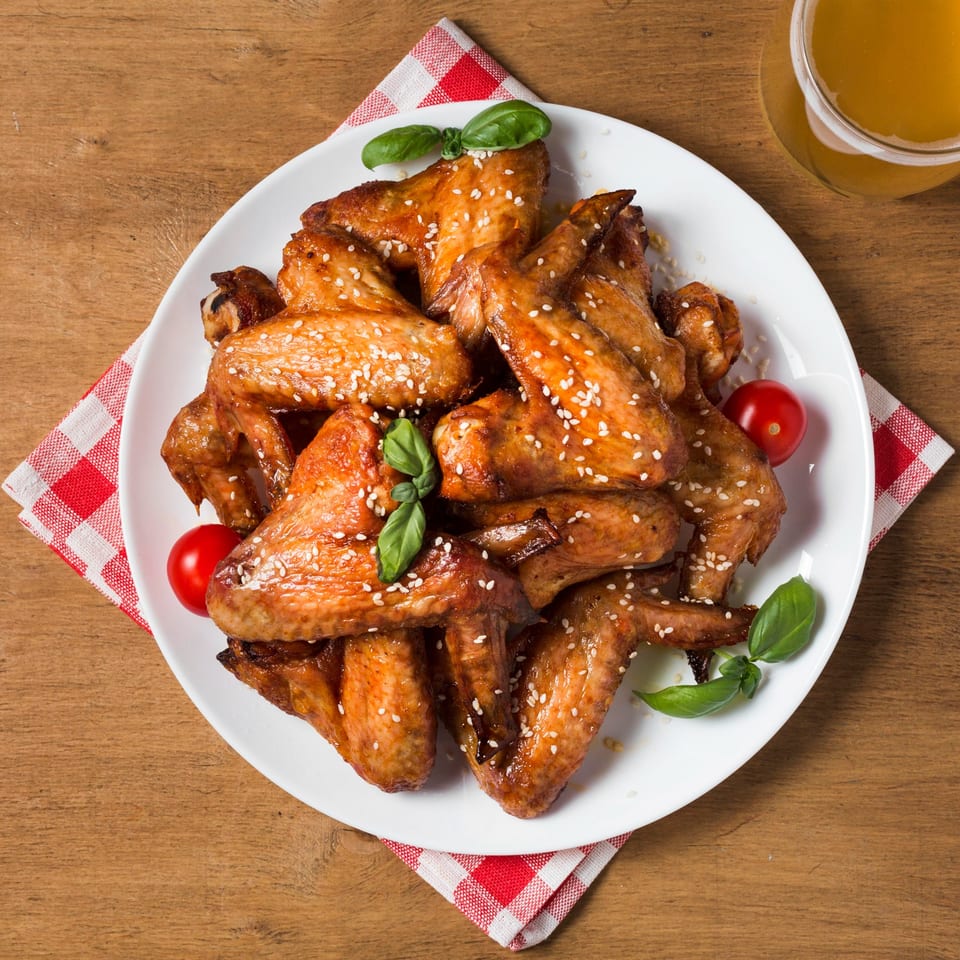 A white plate of sesame chicken wings with basil and cherry tomatoes, next to a glass of beer.