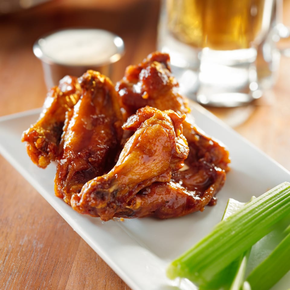 A plate of PieZanos' glazed chicken wings with celery sticks, dipping sauce, and a beer.