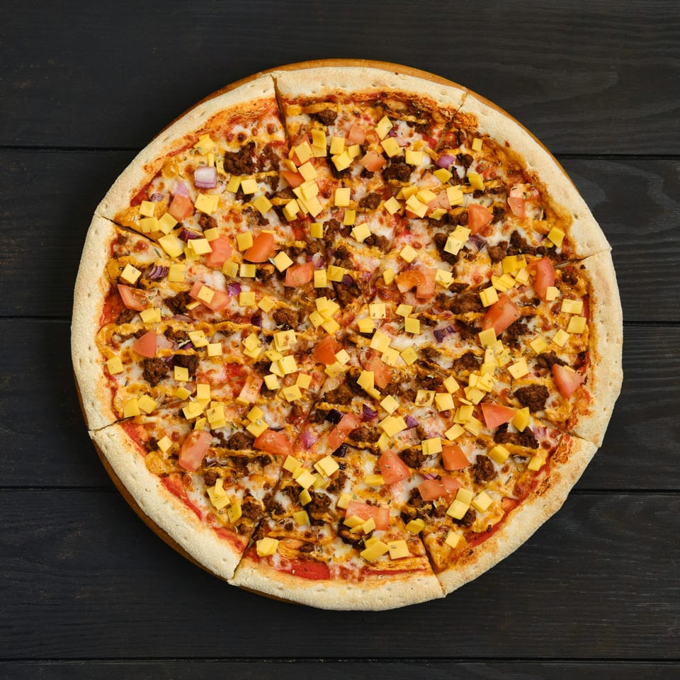 Overhead view of a sliced PieZanos pizza with ground meat, diced tomatoes, and cheese cubes.